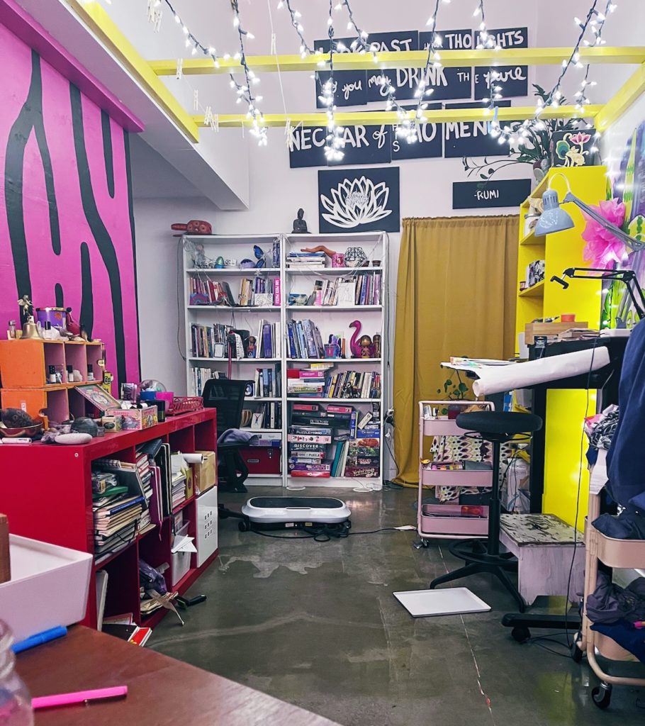 A messy art space, with a hot pink wall on the left, with a shelving unit bursting with stuff against it. Larger white bookshelves on the back wall, and an art table with a pink cart full of art supplies beside it. Above are yellow beams across the ceiling with twinkle lights on them. 